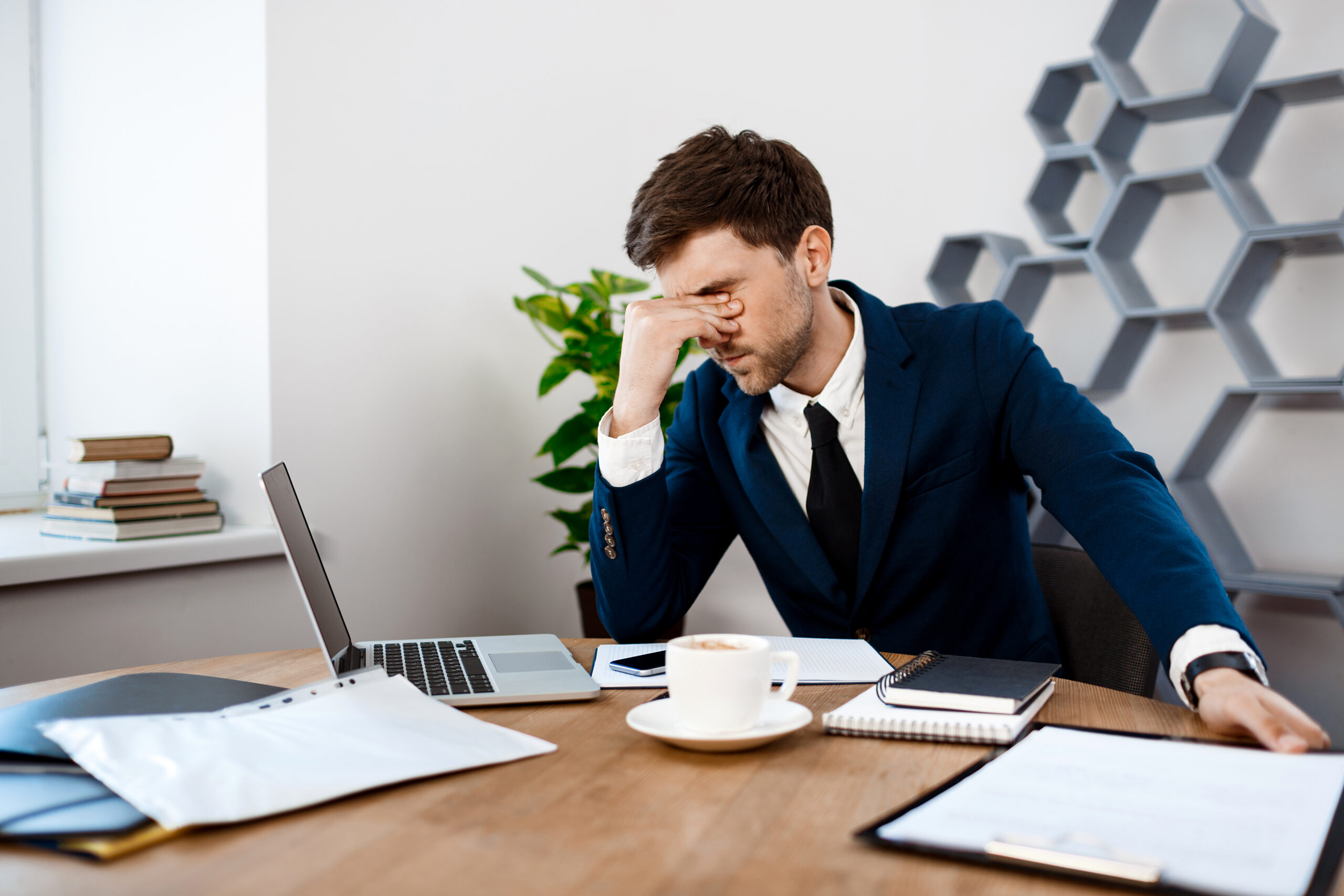 Upset young businessman in suit sitting at workplace, office background.